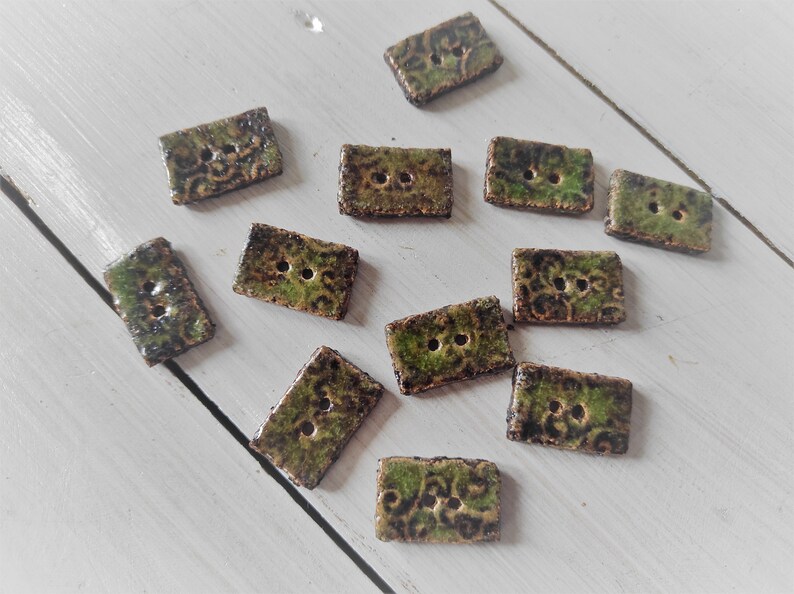 May include: A collection of rectangular ceramic buttons with two holes each. The buttons have a mottled green and brown glaze with a textured, patterned surface. They are scattered on a white wooden surface.