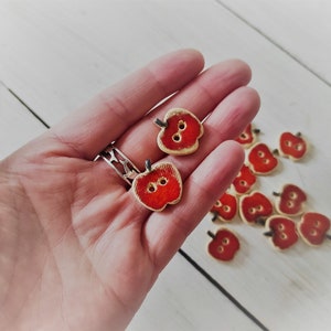 May include: A collection of red and white wooden apple-shaped buttons. The buttons have two holes each and are ideal for sewing or crafting projects.
