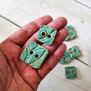 May include: Hand holding several square ceramic buttons. The buttons are a turquoise color with a textured, patterned design and two holes for sewing. The buttons are on a white wooden surface.