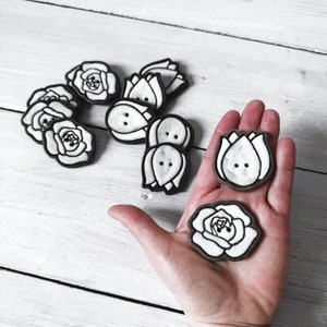 May include: A collection of white ceramic buttons with floral designs. Some buttons are shaped like roses, while others are shaped like tulips. The buttons are all hand-painted with black outlines.