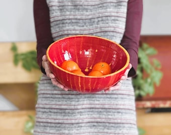 Puede incluir: Un cuenco de cerámica rojo con un glaseado amarillo, lleno de cuatro frutas de color naranja. El cuenco está siendo sostenido por una persona que lleva un suéter a rayas.