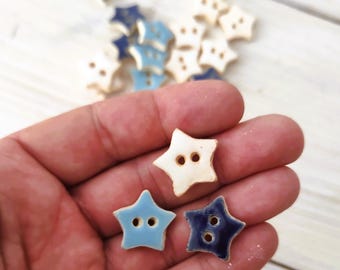 May include: A collection of star-shaped ceramic buttons in various shades of blue and white. The buttons have two holes for sewing and are arranged on a light-colored surface. The buttons are a craft supply.