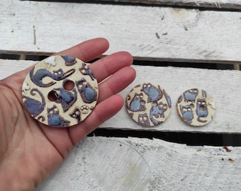 Puede incluir: Tres botones de cerámica con fondo blanco y un patrón de gato azul y negro. Los botones tienen dos agujeros cada uno.