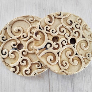 May include: Two cream-colored ceramic buttons with a swirling pattern. The buttons have a two-hole design.