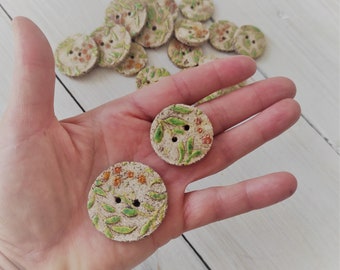 Könnte beinhalten: Eine Sammlung handgemachter Keramikknöpfe mit Blumenmuster. Die Knöpfe sind weiß mit grünen Blättern und orangefarbenen Blüten.