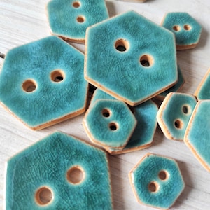 Op de afbeelding: Een verzameling teal keramische knopen met een zeshoekige vorm en twee gaten. De knopen hebben een getextureerd oppervlak en een lichtbruine rand.