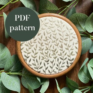 Puede incluir: Un aro de bordado de madera con un fondo de tela blanca con un patrón de hojas verdes y marrones. El texto "PDF pattern" está escrito en blanco sobre un círculo verde oscuro.