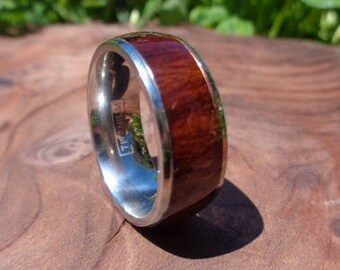 May include: A silver ring with a wide band of reddish-brown wood inlay. The ring is set against a natural wood surface, with a blurred background of green foliage and a white flower.
