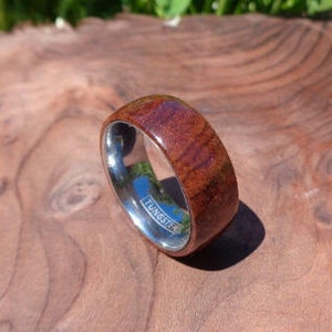 May include: A brown wood and silver tungsten ring. The ring has a polished silver band with the word "TUNGSTEN" engraved on it. The wood portion of the ring has a reddish-brown color and a natural wood grain pattern.