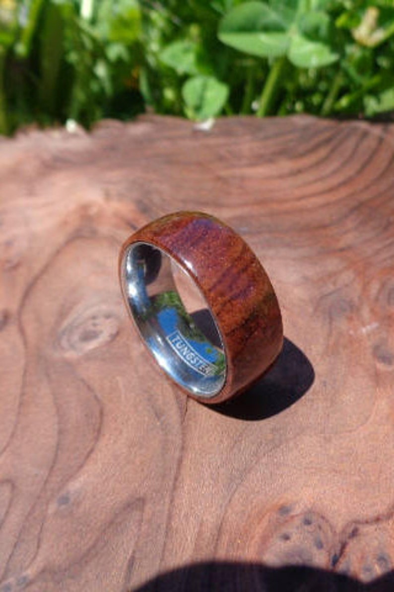May include: A brown wood and silver tungsten ring. The ring has a polished silver band with the word "TUNGSTEN" engraved on it. The wood portion of the ring has a reddish-brown color and a natural wood grain pattern.