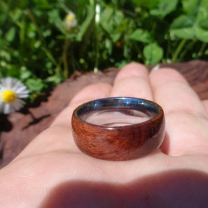 May include: A wooden ring with a dark brown wood grain and a silver metal band. The ring is on a person's finger.