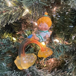 Könnte beinhalten: Vintage Christbaumschmuck mit einer cherubischen Figur mit orangefarbenem Haar und einem grün-weißen Outfit, die auf einem Regenbogen sitzt. Der Regenbogen ist an zwei gelben wolkenförmigen Basen befestigt. Der Schmuck hängt an einem grünen Weihnachtsbaum.