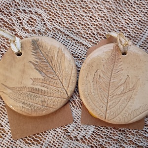 May include: Two round, beige ceramic ornaments with a fern leaf imprint. Each ornament has a small hole at the top with a gold and white ribbon for hanging. The ornaments are on a textured, light brown surface.