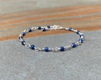 May include: A delicate bracelet featuring small, round, deep blue beads strung on a silver chain. The bracelet is designed with a simple, elegant style, perfect for everyday wear. The beads are evenly spaced along the chain, creating a consistent pattern.