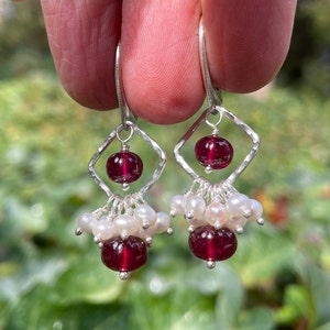 May include: A pair of silver earrings with a red gemstone and white pearl accents. The earrings feature a square frame with a red gemstone in the center and a cluster of white pearls hanging below.