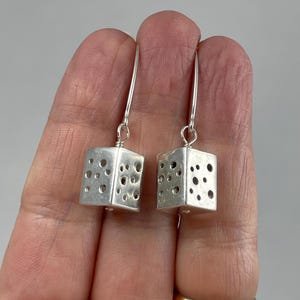 May include: A pair of silver dangle earrings with a geometric cube design. Each cube has a pattern of small circles cut out of the surface.
