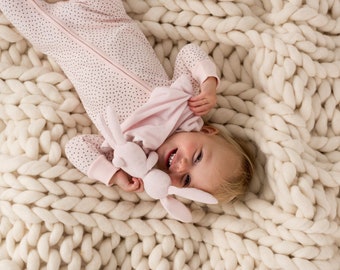May include: A baby wearing a pink and white polka dot sleepsuit is lying on a white chunky knit blanket. The baby is holding a pink plush bunny toy and smiling.