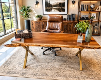 May include: A live edge wooden desk with gold-colored metal legs. The desk is in a home office setting, with a leather chair, a wooden cabinet, and a bookshelf. A framed landscape painting hangs on the wall.