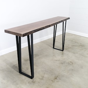 May include: A long, dark wood table with a live edge top and black metal hairpin legs. The table is standing on a concrete floor.