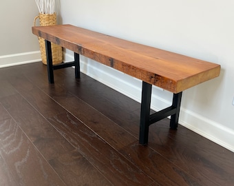 May include: A wooden bench with a natural wood finish and black metal legs. The bench is sitting on a dark wood floor.