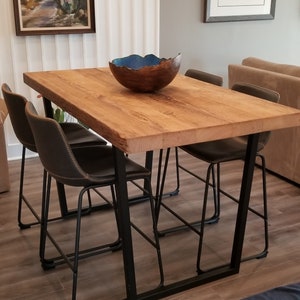 May include: A rustic wooden dining table with a live edge, supported by black metal legs. Four dark gray bar stools with stitched detailing are positioned around the table. A decorative bowl with a blue interior sits on the table.