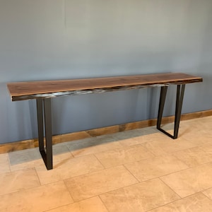 Pu&ograve; includere: Un lungo tavolo console in legno scuro con un piano a bordo vivo e gambe in metallo nero. Il tavolo &egrave; su un pavimento piastrellato.
