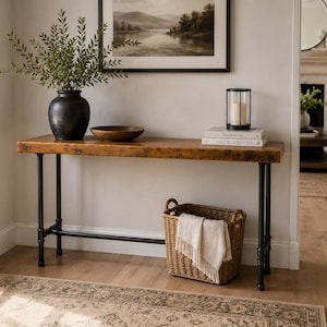 May include: A rustic wooden console table with black metal legs. A black vase with greenery, a wooden bowl, and a candle lantern sit on top. A woven basket with a cream-colored throw is below. A framed landscape painting hangs above.