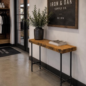 May include: A rustic wooden console table with black metal pipe legs. A dark gray ceramic pot with greenery sits on the table, along with a stack of books. A sign on the wall reads "IRON & OAK SUPPLY CO. EST. 2016."
