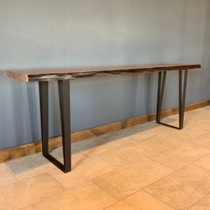 Pu&ograve; includere: Un tavolo consolle lungo in legno scuro con un piano a bordo vivo e gambe in metallo nero. Il tavolo ha un aspetto rustico e industriale e sarebbe perfetto per un corridoio o un ingresso.