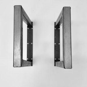 May include: Two metal table legs with a rectangular shape and a welded construction. The legs are made of a dark gray metal and have a matte finish.