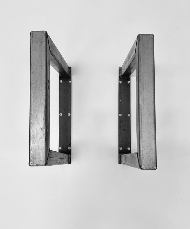 May include: Two metal table legs with a rectangular shape and a welded construction. The legs are made of a dark gray metal and have a matte finish.