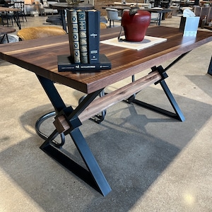 May include: A dark wood table with a black metal X-shaped base. The table has a natural wood finish and is accented with a wooden crossbar. The table is in a showroom setting.