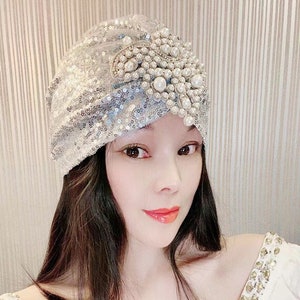 Puede incluir: Un turbante de lentejuelas plateadas con un gran adorno de perlas y pedrería.