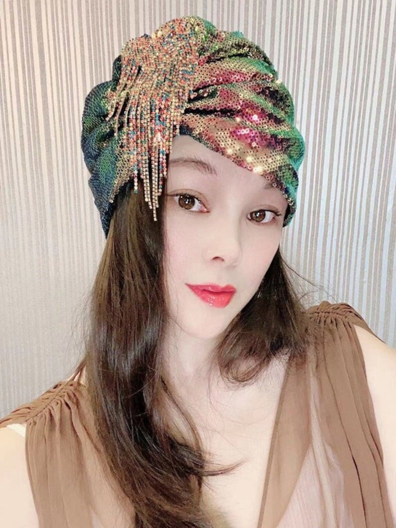May include: A sequined turban in shades of green, pink, and gold, adorned with a sparkling, dangling fringe. The head covering is a glamorous accessory, perfect for special occasions or adding a touch of flair to any outfit.