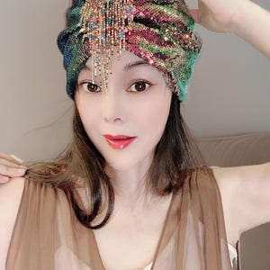 May include: A sequined turban in shades of green, blue, and pink, adorned with a beaded fringe. The head covering is a stylish accessory, perfect for adding a touch of glamour to any outfit.
