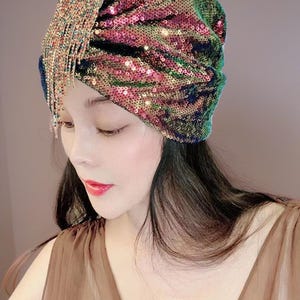 May include: A sequined turban in shades of green, pink, and gold. The turban features a draped design and a sparkling fringe detail on the side. The head covering is a glamorous accessory.