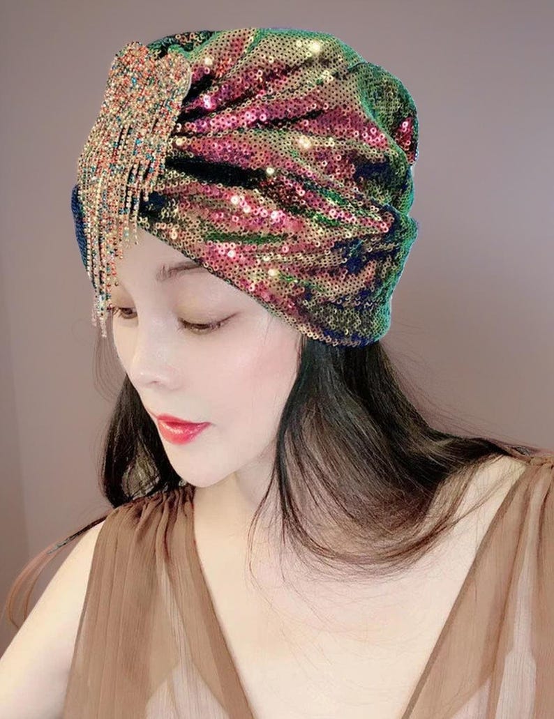 May include: A sequined turban in shades of green, pink, and gold. The turban features a draped design and a sparkling fringe detail on the side. The head covering is a glamorous accessory.