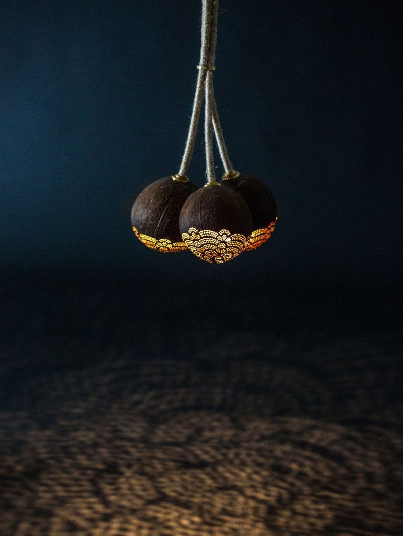 May include: A three-light pendant lamp made from coconut shells with intricate cut-out designs. The shells are a natural brown color and the light shines through the cut-outs, creating a warm glow.