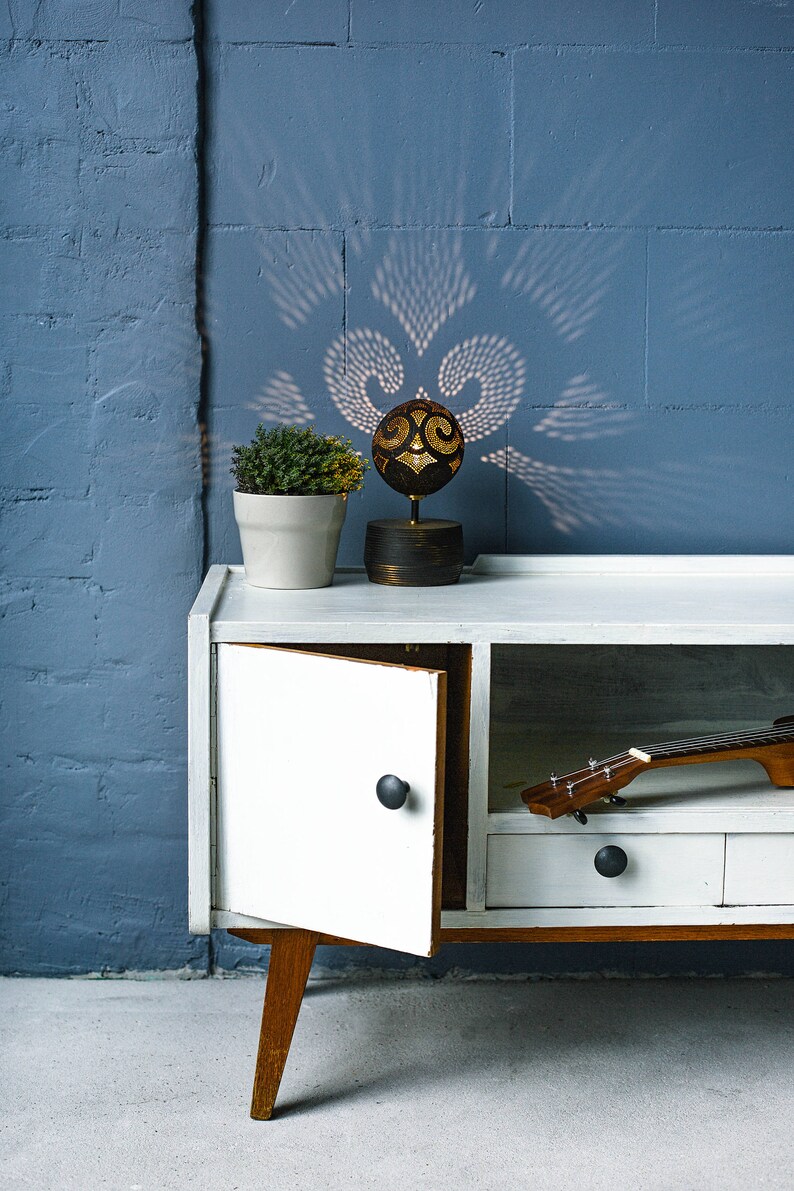 Pode incluir: Um arm&aacute;rio branco com uma base de madeira e uma pequena planta em um vaso branco no topo. O arm&aacute;rio tem uma gaveta e uma porta que est&aacute; aberta, revelando um ukulele dentro. Uma l&acirc;mpada decorativa feita de uma casca de coco com um design entalhado fica no arm&aacute;rio. A l&acirc;mpada projeta uma sombra na parede atr&aacute;s dela.