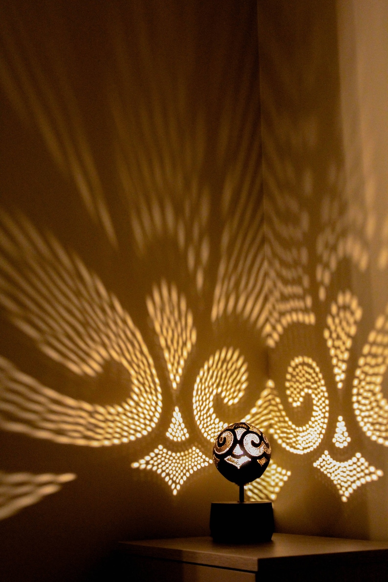 Pode incluir: Uma l&acirc;mpada de coco esculpida projeta padr&otilde;es intrincados de luz na parede, criando uma atmosfera quente e acolhedora. A l&acirc;mpada &eacute; feita de casca de coco natural e apresenta um design &uacute;nico.