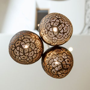 May include: Three round, brown, coconut shell light fixtures with intricate cut-out designs. The lights are hanging from the ceiling and are illuminated.
