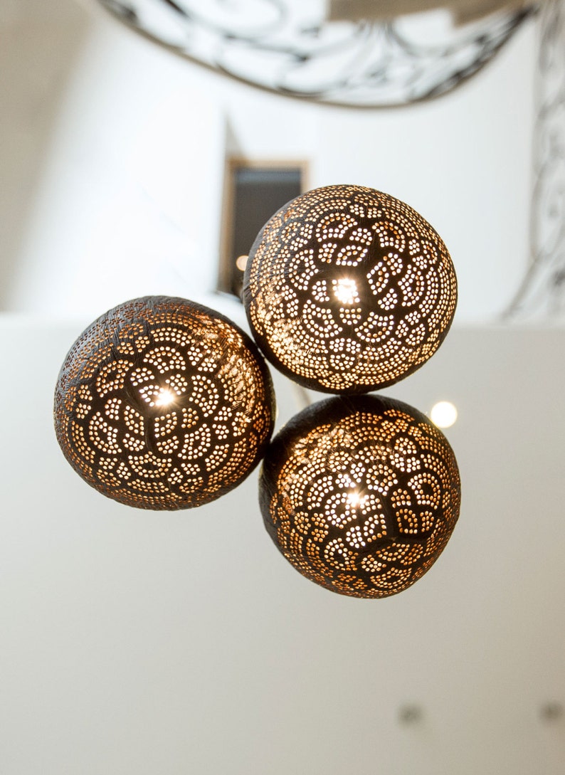May include: Three round, brown, coconut shell light fixtures with intricate cut-out designs. The lights are hanging from the ceiling and are illuminated.