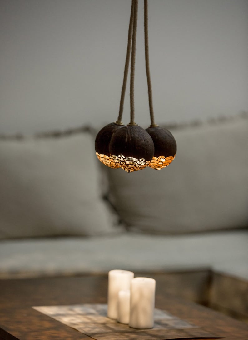 May include: Three coconut shell light fixtures hang from a rope, with intricate cutouts around the bottom of each shell. The lights are illuminated, casting a warm glow. Three white candles are on a wooden table in the foreground.