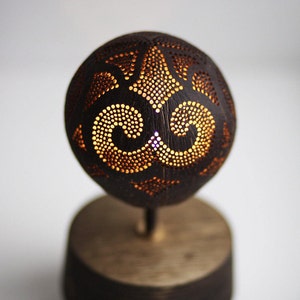 Pode incluir: Uma pequena l&acirc;mpada de mesa feita de uma casca de coco com um design entalhado. A casca &eacute; iluminada por dentro, criando um brilho quente atrav&eacute;s do padr&atilde;o intrincado.