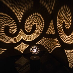 Pode incluir: Uma pequena l&acirc;mpada redonda decorativa com um design entalhado projeta padr&otilde;es intrincados de luz em uma parede escura. A l&acirc;mpada &eacute; feita de um material claro e tem um brilho quente e acolhedor.