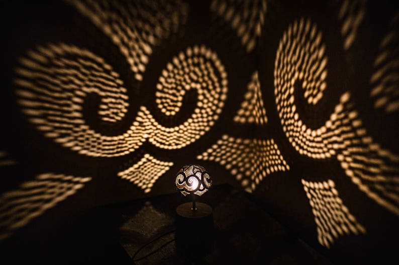 Pode incluir: Uma pequena l&acirc;mpada redonda decorativa com um design entalhado projeta padr&otilde;es intrincados de luz em uma parede escura. A l&acirc;mpada &eacute; feita de um material claro e tem um brilho quente e acolhedor.
