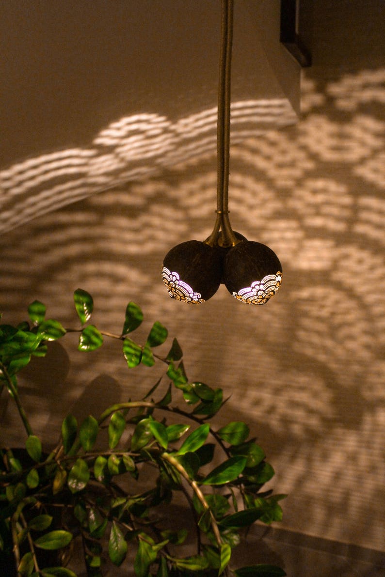 May include: A hanging light fixture made from two carved coconut shells with intricate designs. The shells are illuminated from within, casting a warm glow.