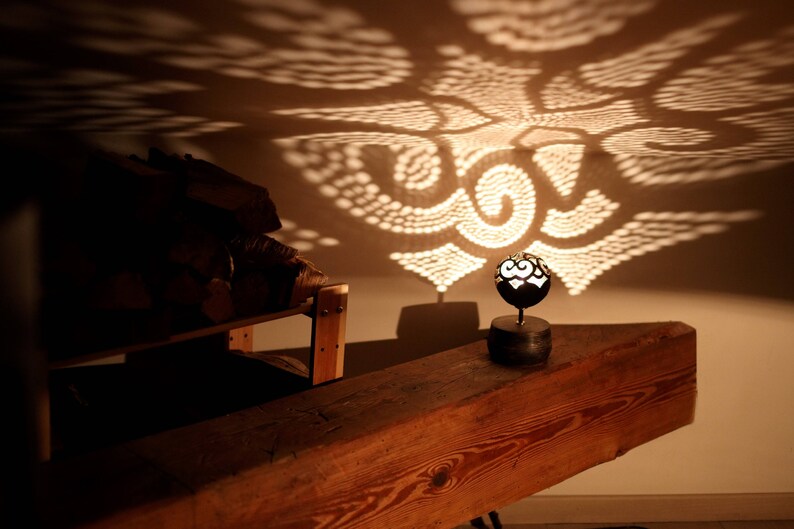 Pode incluir: Uma l&acirc;mpada de metal decorativa com um design geom&eacute;trico projeta uma sombra estampada sobre uma superf&iacute;cie de madeira. A l&acirc;mpada est&aacute; sobre uma base pequena e tem uma l&acirc;mpada dentro.
