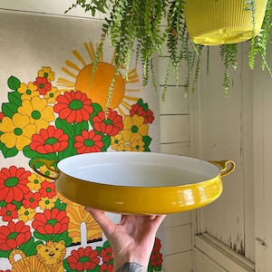 May include: A vintage, round, yellow enamel pan with a white interior is held up by a hand. The pan has two small handles. The background includes a floral tapestry with a sun design and hanging plants.