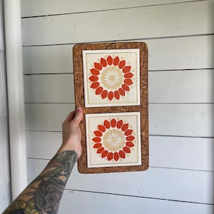 May include: A decorative wall hanging with two square tiles, each displaying a floral design. The tiles have a cream background with red and gold flower patterns. They are set in a rectangular, textured, brown frame.